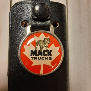 Op de afbeelding: Een zwarte leren sleutelhanger met een zilveren sleutelring en een ronde rode en witte hanger. De hanger heeft een bulldog, een esdoornblad en de tekst "Mack Trucks".