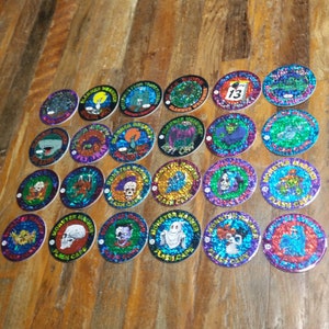 May include: A collection of 25 round, holographic, silver-coloured tokens with various monster and creature designs. Each token features a different image, including a ghost, a skull, a vampire, and a werewolf. The tokens are arranged in a grid pattern on a wooden surface.
