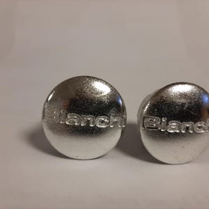 May include: Two silver-coloured, round metal objects with the word "Bianchi" embossed on each. The objects have a shiny, reflective surface and are set against a plain background. The items appear to be small, possibly decorative or functional.