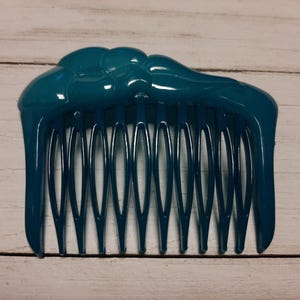 May include: A teal blue plastic hair comb with a decorative top. The comb has a wavy design and 10 teeth.