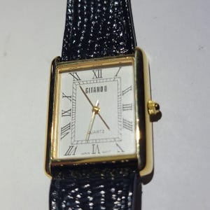 May include: A gold rectangular wristwatch with a white face and black Roman numerals. The watch face reads "GITANO" and "QUARTZ". The watch has a black textured leather strap.