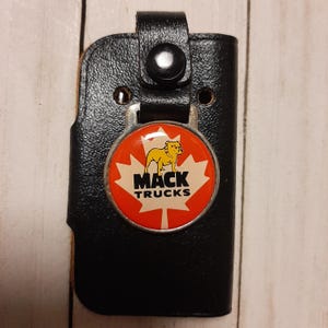 Op de afbeelding: Zwarte leren sleutelhouder met een rond embleem met het Mack Trucks-logo. Het embleem heeft een rood en wit esdoornbladontwerp met een gele bulldog en de woorden "MACK TRUCKS". De sleutelhouder heeft een drukknoopsluiting.