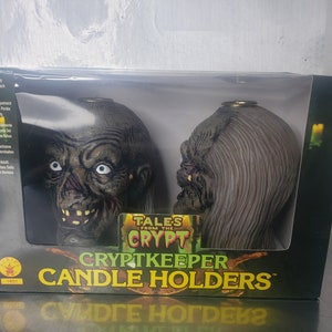 Könnte beinhalten: Zwei grüne und graue Cryptkeeper-Kerzenhalter aus Tales from the Crypt. Die Kerzenhalter sind in Form des Kopfes des Cryptkeepers gestaltet. Auf der Schachtel steht "Cryptkeeper Candle Holders".