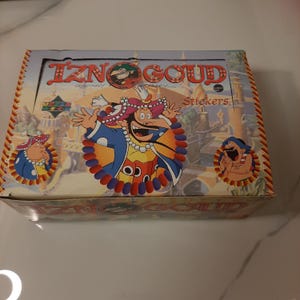 May include: A box of Iznogoud stickers featuring a cartoon character with a large moustache and a red and blue turban. The box is yellow and orange with the text "Iznogoud" in large black letters.