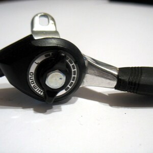 Pode incluir: Um desviador de bicicleta Shimano preto e prateado com uma pega de borracha preta. O desviador tem um dial com números.