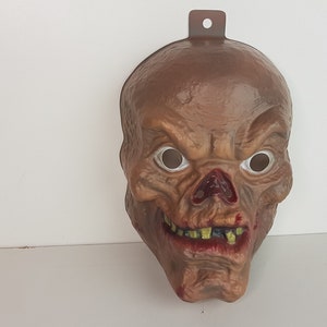 Op de afbeelding: Een bruin plastic Halloween-masker van een schedel met een bloederige neus en tanden. Het masker heeft witte ogen en een gat bovenaan om op te hangen.