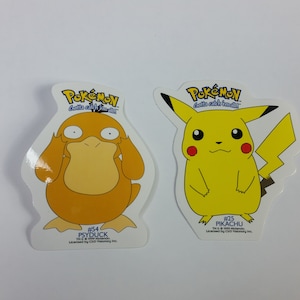 May include: Two Pokemon character stickers, one of Pikachu and one of Psyduck. The stickers are white with a yellow background and feature the Pokemon characters in a cartoon style. The text on the stickers reads "Gotta catch 'em all!" and includes the Pokemon character's name and number.