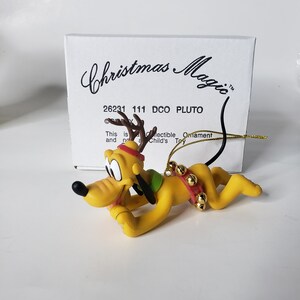 May include: A yellow Pluto figurine wearing a red and green reindeer antler headband and a red and gold jingle bell collar. The figurine is in a crouching position with its head turned to the left. The figurine is on a white background with a box that says "Christmas Magic" and "26231 111 DCO PLUTO".