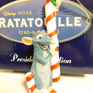 May include: A blue rat character from the Disney Pixar film Ratatouille holding a red and white candy cane ornament. The ornament is labelled "Disney Pixar Ratatouille (rat.a.touille)" and "Presid.tion".