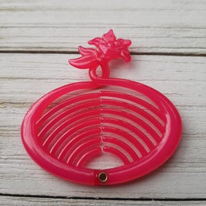 May include: A pink plastic hair comb with a flower design. The comb has a rounded oval shape with a series of ridges on one side. The flower is a stylised design with five petals.