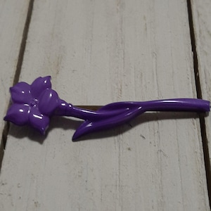 May include: A purple plastic flower with a single stem and leaf. The flower has five petals and is shaped like a lily.