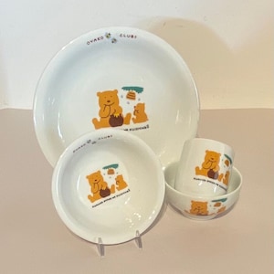 Könnte beinhalten: Ein Set aus weißen Keramikgeschirr mit einem Cartoon-Bären-Design. Das Set enthält einen großen Teller, eine kleinere Schüssel und eine Tasse. Das Design zeigt zwei Bären mit Honigbehältern und Bienen. Der Text "OYAKO CLUB" ist auf dem Geschirr gedruckt.