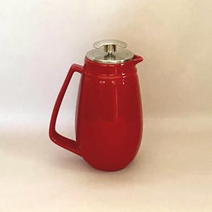 May include: A bright red ceramic French press coffee maker with a glossy finish. It features a sturdy, matching red handle on the side and a metallic silver lid with a central plunger knob. The body has a rounded, bulbous shape with a small spout.