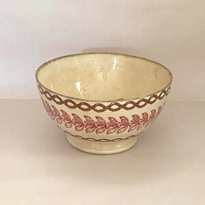 Peut inclure: Un bol en céramique de couleur crème orné d'un motif de corde marron sur le bord et la base, avec une bande centrale de motifs de feuilles rouges. Le bol a une forme arrondie et repose sur un petit pied, présentant une finition émaillée.