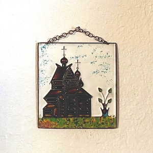 Puede incluir: Un colgante de pared de cerámica con una imagen pintada de una iglesia con un techo verde y un pequeño árbol. La iglesia tiene un exterior de madera marrón y una cruz blanca en la parte superior. El colgante de pared está sujeto a una pared blanca con una cadena.