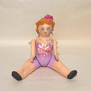 May include: A handmade doll with a painted face, red hair, and a pink bow. The doll wears a lavender leotard with a floral design and has outstretched arms and legs. The doll's feet are painted black.