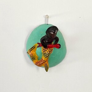 May include: A wall-mounted decorative plaque featuring a hand-painted mermaid with a black torso and a yellow tail with black scale details. The mermaid holds a red fish against a circular, light turquoise background with a metal hanging loop.