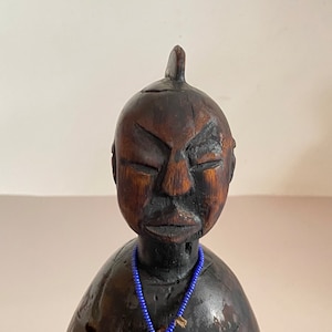 Puede incluir: Escultura de madera marrón oscuro de una persona con los ojos cerrados y una pequeña punta en la parte superior. La escultura tiene un collar de cuentas azules con un colgante. La madera tiene vetas visibles y una textura rugosa.
