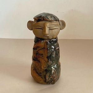 May include: A handcrafted ceramic figurine with a stylized human face and body. The sculpture features a brown and green color palette, with a textured surface. The figure has a hat and is a unique piece of art.