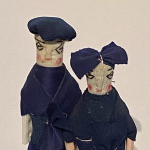 Könnte beinhalten: Zwei handgefertigte Vintage-Puppen. Eine Puppe trägt ein marineblaues Barett und eine Jacke, die andere eine marineblaue Schleife und eine Jacke. Beide Puppen haben bemalte Gesichter mit rosigen Wangen und dunklen Augen. Die Puppen sind aus Stoff und Holz.