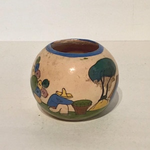 May include: A small, round, ceramic vase with a blue rim and a hand-painted scene of a person in a blue shirt and yellow hat picking fruit from a tree. The vase has a light brown background with green, yellow, and blue accents.
