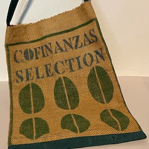 May include: A brown burlap tote bag with a green trim and a black strap. The bag has a green and white coffee bean design and the text "COFINANZAS SELECTION" printed on the front.
