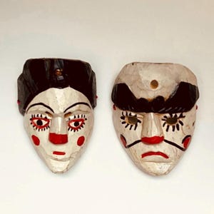 Peut inclure: Deux masques en bois peints à la main avec des détails noirs, blancs et rouges. Les masques ont des traits exagérés, notamment de grands yeux, des nez et des bouches. Un masque a un chapeau noir et l'autre une moustache noire.