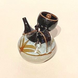 May include: A small, handmade ceramic teapot with a distinctive design. The teapot is glazed in shades of brown, white, and blue, featuring a stylised leaf pattern. The teapot has a small, round lid with a knob on top.