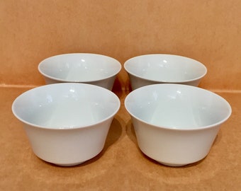 May include: Four white ceramic bowls, arranged in a square formation on a brown surface.