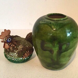 May include: A green ceramic vase with a dark green glaze and a brown rim. The vase has a round body and a wide opening. A small, beaded, brown and green chicken figurine is in the foreground.