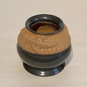 May include: A small, round, brown clay pot with a black glaze on the bottom half. The pot has a decorative design carved into the top half.