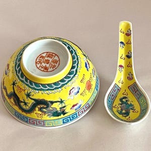 以下が含まれることがあります： 龍のデザインが施された黄色の磁器のボウルとスプーン。ボウルは緑と青の縁取りがあり、スプーンは緑と青の柄。両方の作品には底に中国語の文字が書かれている。