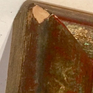 May include: Close-up of a wooden frame with a chipped corner. The frame is painted in shades of red, brown, and gold. The frame is likely for a painting or artwork.