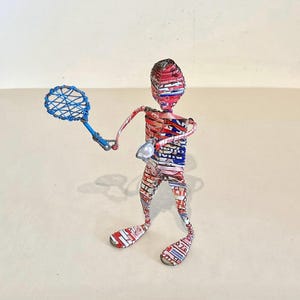 Könnte beinhalten: Eine Drahtfigur, die eine Person beim Tennisspielen darstellt. Die Skulptur ist aus recycelten Materialien hergestellt, darunter Aluminiumdosen. Die Person trägt ein rot-weiß-blaues Outfit und hält einen blauen Tennisschläger. Die Skulptur steht auf einer weißen Oberfläche.