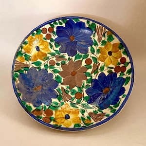 May include: A round, hand-painted ceramic plate with a floral design. The plate features large blue, yellow, and brown flowers with green leaves and brown berries against a white background. The rim is a dark blue.