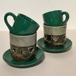 May include: Two green ceramic coffee cups with matching saucers. The cups are stacked on top of each other and have a white and black design with gold accents.
