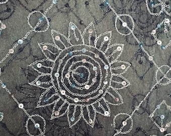 Puede incluir: Primer plano de una tela negra con lentejuelas plateadas y azules dispuestas en un patrón circular. Las lentejuelas están cosidas en un diseño floral con un círculo más pequeño en el centro.
