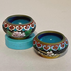 Puede incluir: Dos pequeños cuencos redondos de cloisonné con intrincados diseños florales en rojo, blanco y verde sobre un fondo negro. Los cuencos tienen un interior turquesa y se asientan sobre una base turquesa. Los bordes están ribeteados en dorado.