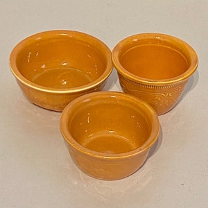 May include: Three ceramic bowls in various sizes, all in a warm orange color. The bowls are glazed and have a smooth, slightly textured surface.