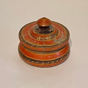 May include: A small, round wooden box with a lid. The box is painted in shades of orange and black with intricate designs. The lid has a knob on top.