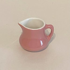 以下が含まれることがあります： 白い内側と取っ手のある小さなピンクのセラミック製のクリーマーです。