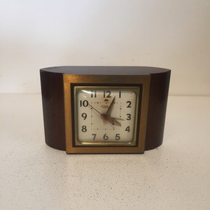 以下が含まれることがあります： 金色の正方形の文字盤が付いた、ヴィンテージの木製時計。 時計の文字盤には黒い数字と針があり、「Selva」のブランド名が見えます。 時計は濃い茶色の木製のベースに置かれています。