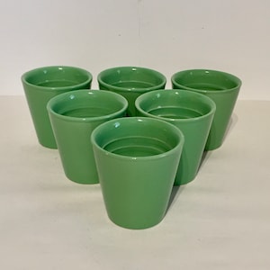 Könnte beinhalten: Sechs grüne Keramikbecher, die in einem Kreis auf einer weißen Oberfläche angeordnet sind. Die Becher sind alle gleich groß und gleich geformt und haben eine glatte, glänzende Oberfläche.