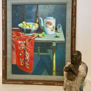 May include: A framed painting of a still life with a teapot, fruit, and a musical instrument. The painting is set on a table with a red cloth draped over the side. A small ceramic statue of a Buddha is in the foreground.