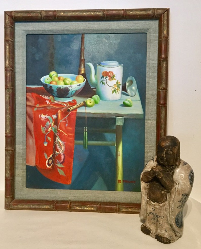 May include: A framed painting of a still life with a teapot, fruit, and a musical instrument. The painting is set on a table with a red cloth draped over the side. A small ceramic statue of a Buddha is in the foreground.