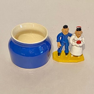 May include: A cobalt blue ceramic jar with a cream-colored interior sits next to a small figurine of two people. The figurine depicts a person in a blue jumpsuit and another in a white coat, standing on a yellow base.