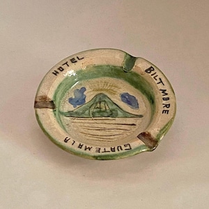 Puede incluir: Cenicero de cerámica hecho a mano en verde y crema. Presenta una escena de montaña pintada y las palabras "HOTEL BILTMORE GUATEMALA". El cenicero tiene una pequeña muesca para apoyar los cigarrillos. Diámetro aproximado de 10 cm.