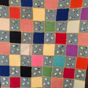 May include: A colorful knitted patchwork quilt with a checkered pattern. Each square is a different color and features a small embroidered design of leaves.