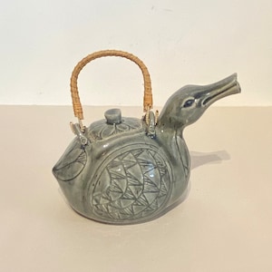 May include: A ceramic teapot shaped like a duck with a woven handle. The teapot is a light blue-green color with a geometric pattern.