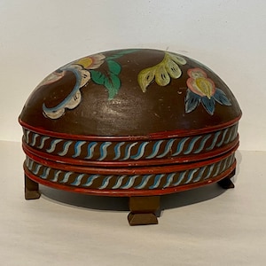 Puede incluir: Una caja redonda de madera con una tapa abovedada pintada con diseños florales coloridos. La caja tiene una base marrón con bandas pintadas de rojo y azul y cuatro patas cortas.